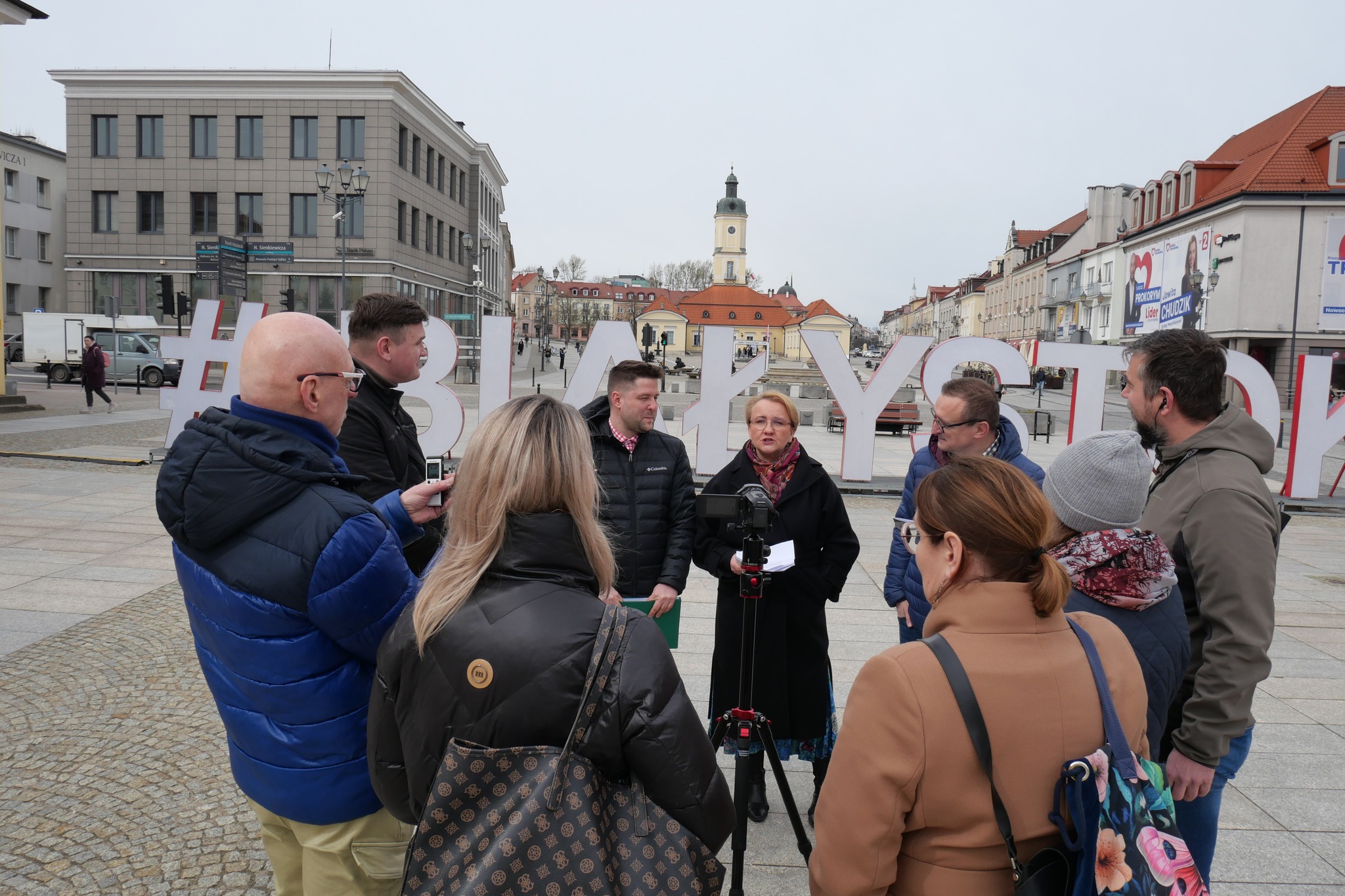 Kilka osób stoi na Rynku Kościuszki w centrum Białegostoku. W tle widać ratusz miejski. Jest to konferencja prasowa, trzy osoby wypowiadają się do mikrofonu. Dziennikarze i dziennikarki stoją w pierwszym planie.